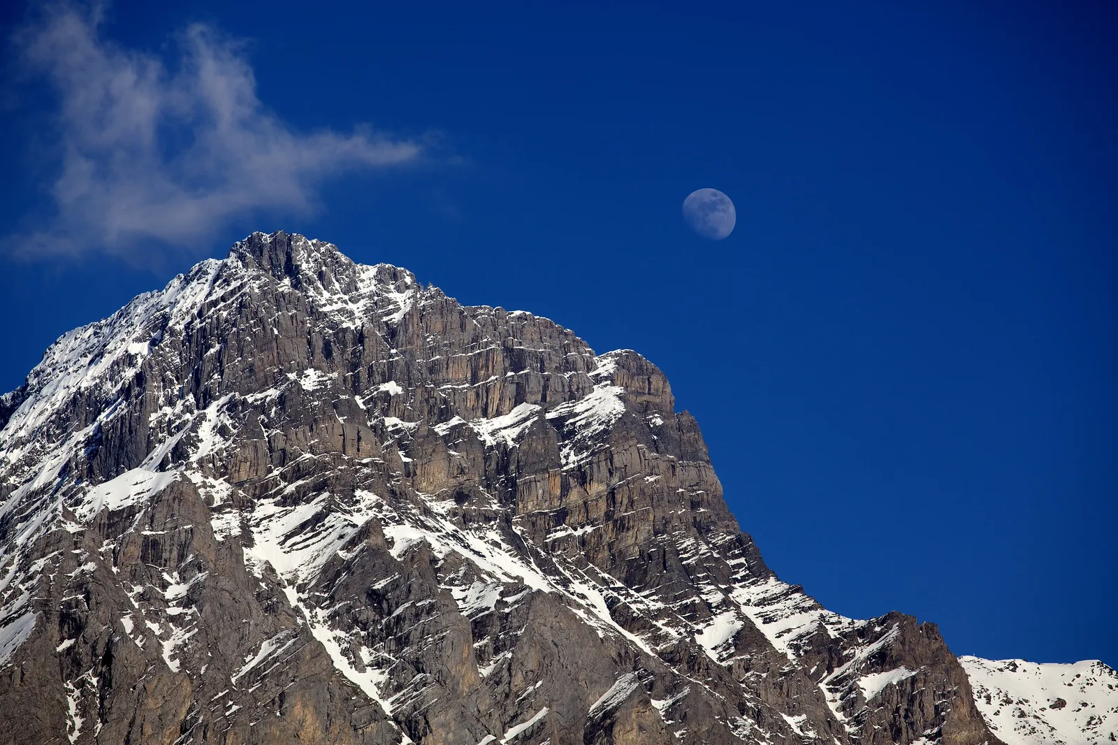 A pale gibbous moon in a deep blue daytime sky above a steep rocky mountain peak streaked with snow, with a thin white cloud at the upper left