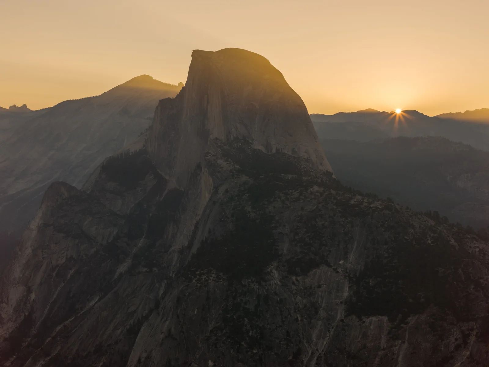 A large granite dome in shadow at sunrise with the sun just clearing a distant ridge and several mountain ridges fading into orange haze behind it
