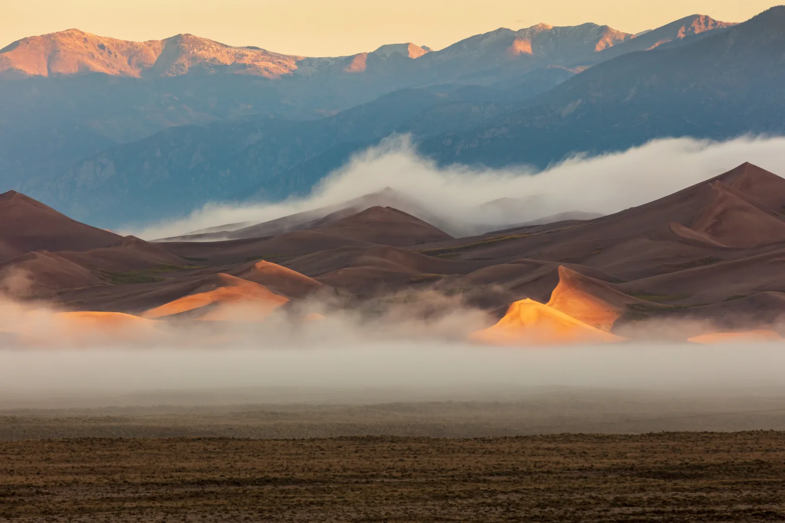 Sand dunes lit orange by low sun, with a band of fog in front and blue mountains with pink peaks behind
