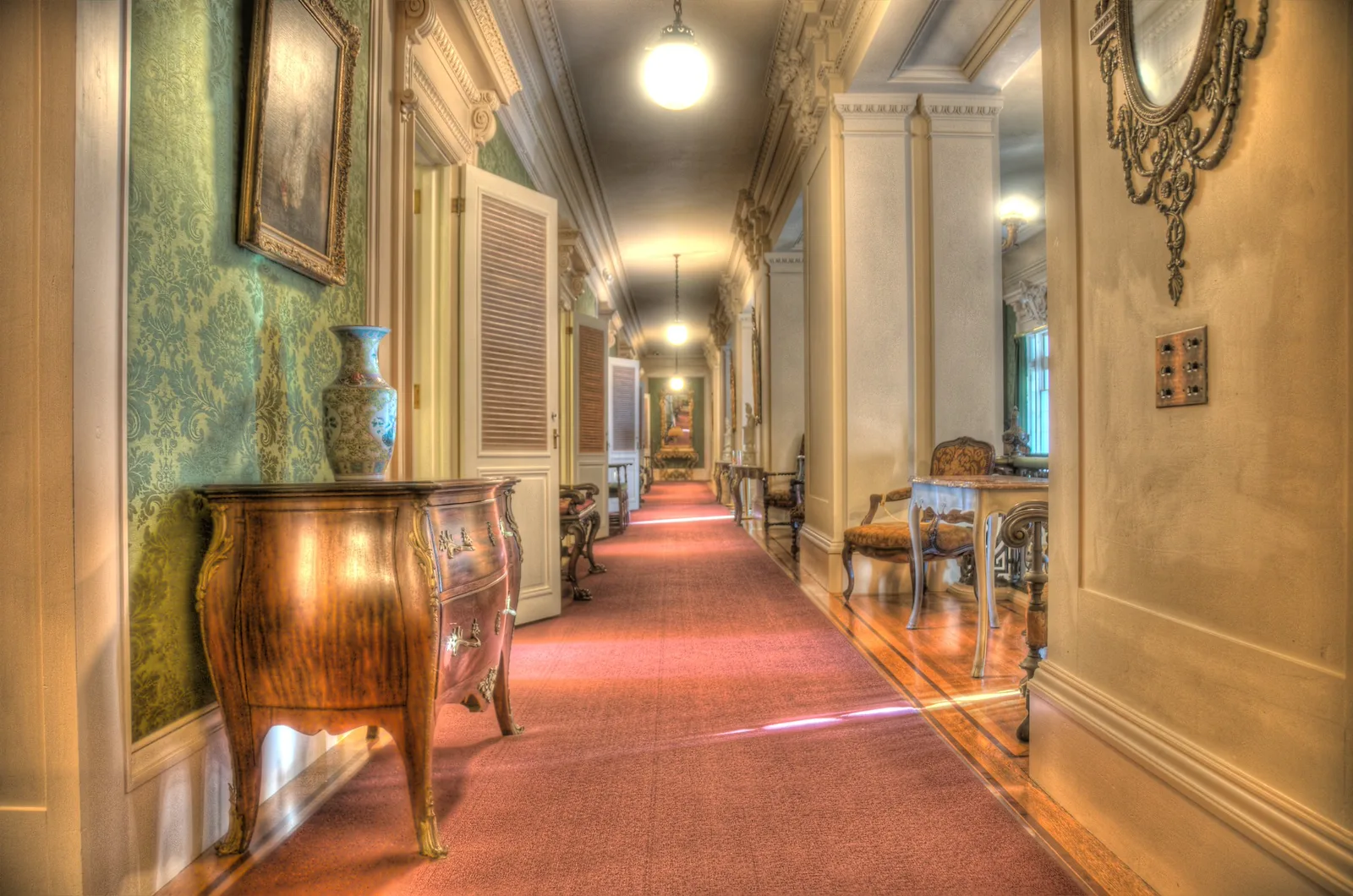 A long carpeted hallway in a historic house with a framed picture hung above a wooden chest on the left wall and an ornate mirror on the right
