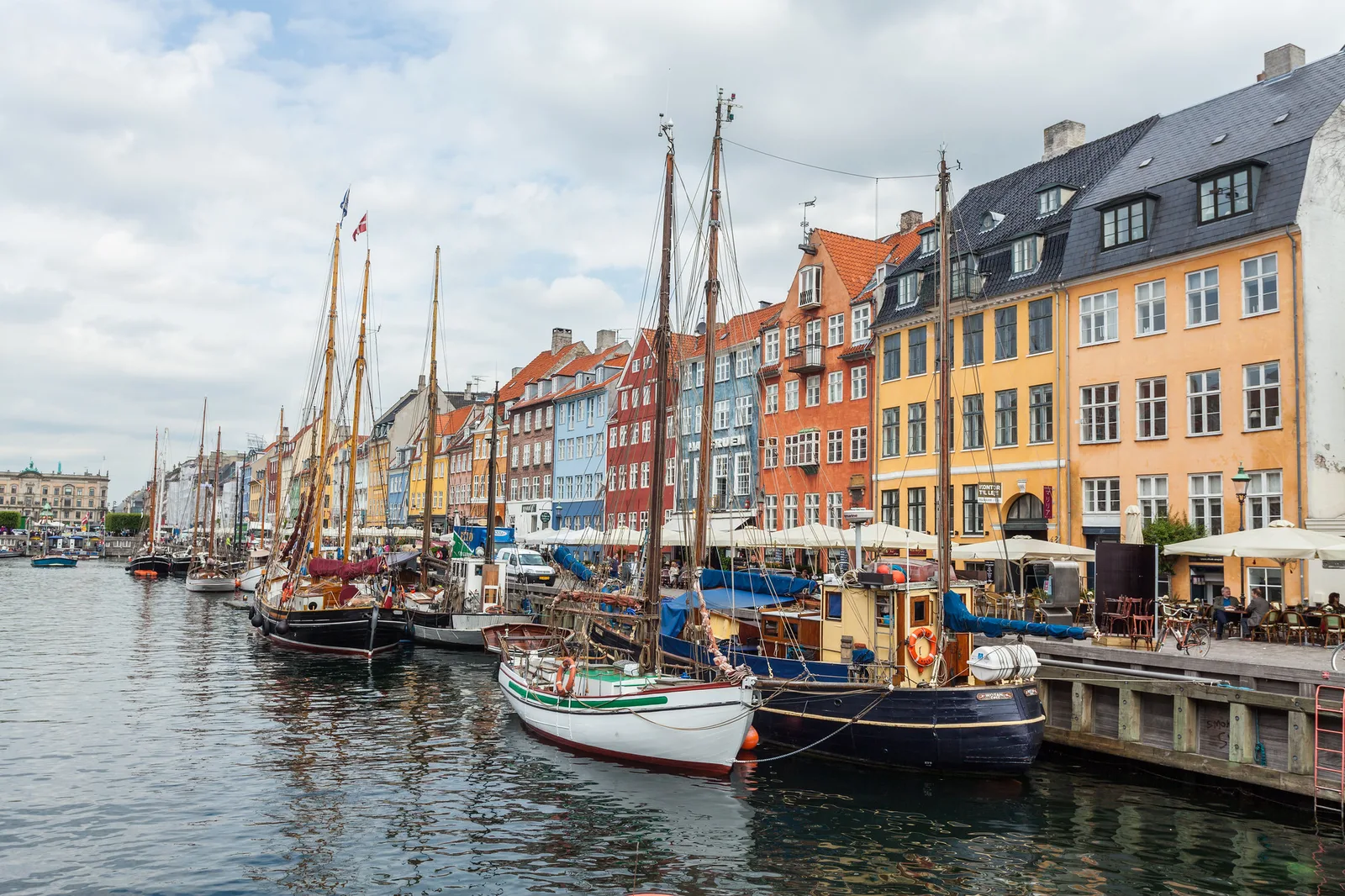 Wooden sailing boats moored along a canal quay lined with tall yellow, orange and blue townhouses and cafe umbrellas under a cloudy sky