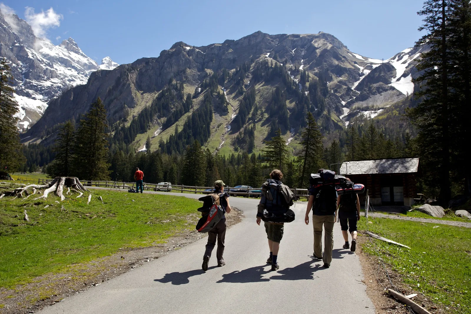 Four hikers with large backpacks seen from behind, walking along a road toward a wall of rocky mountains with snow patches, with a small hut and trees beside them
