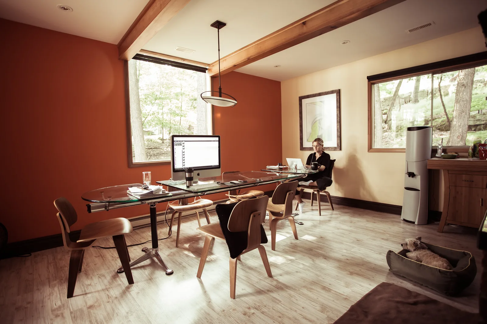 A warm-toned home office with a desktop computer on a glass table, wooden chairs around it and a person working at a second desk in the background