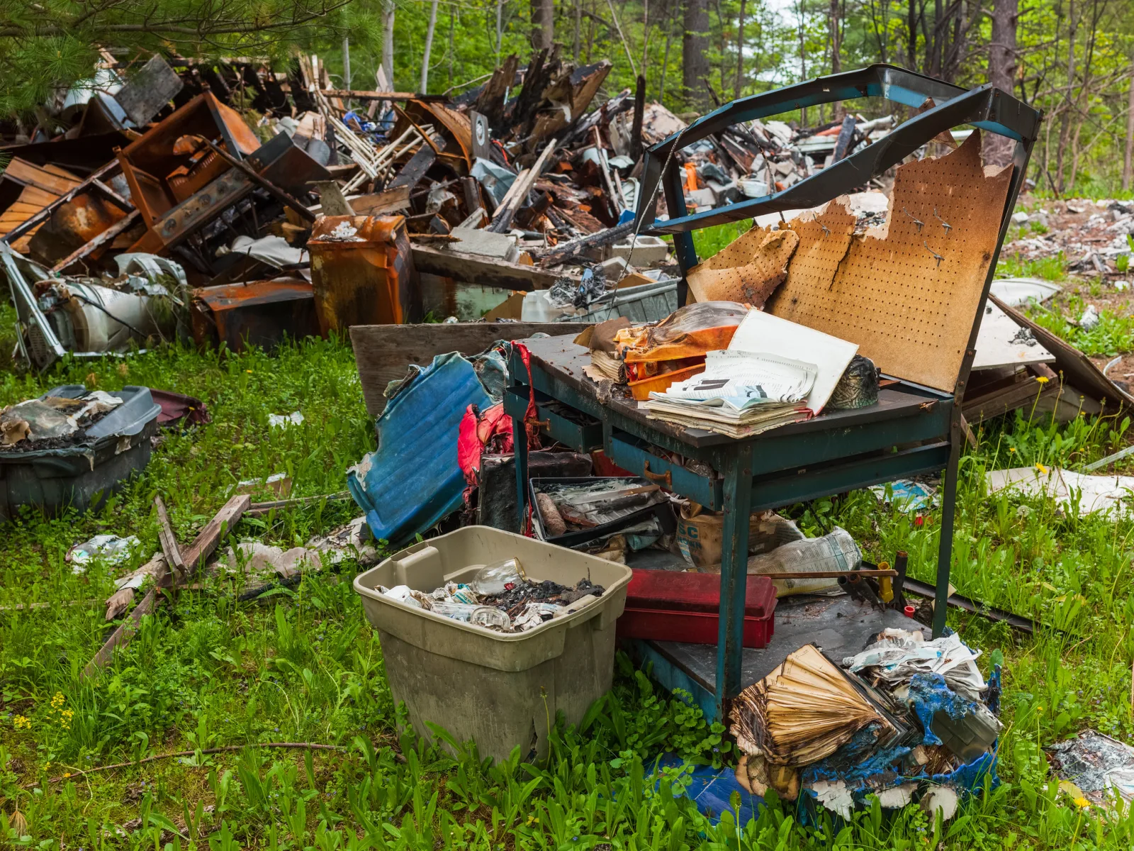 A heap of broken, charred furniture and household debris on grass, including a table, a plastic storage bin and scattered papers