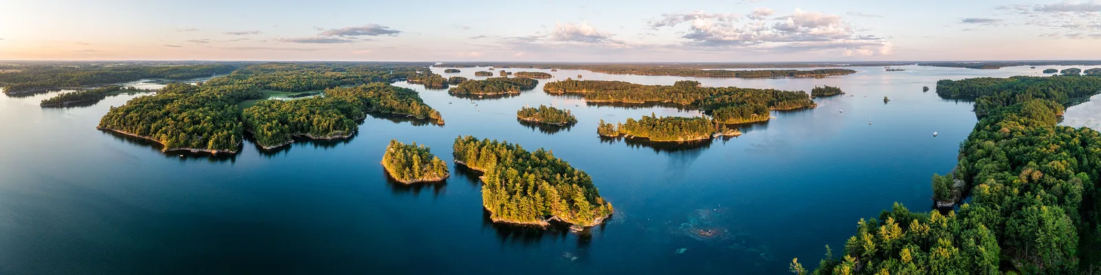 Very wide aerial panorama of wooded islands scattered across calm blue water at dusk under a pale sky