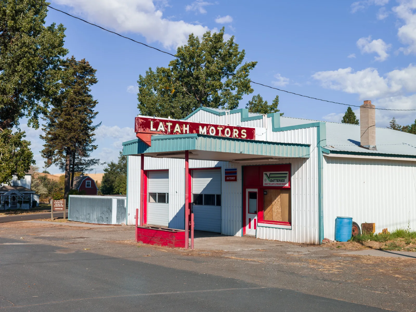 A small white garage with a red Latah Motors sign and red trim, photographed from the roadside in bright sun under a blue sky with scattered clouds