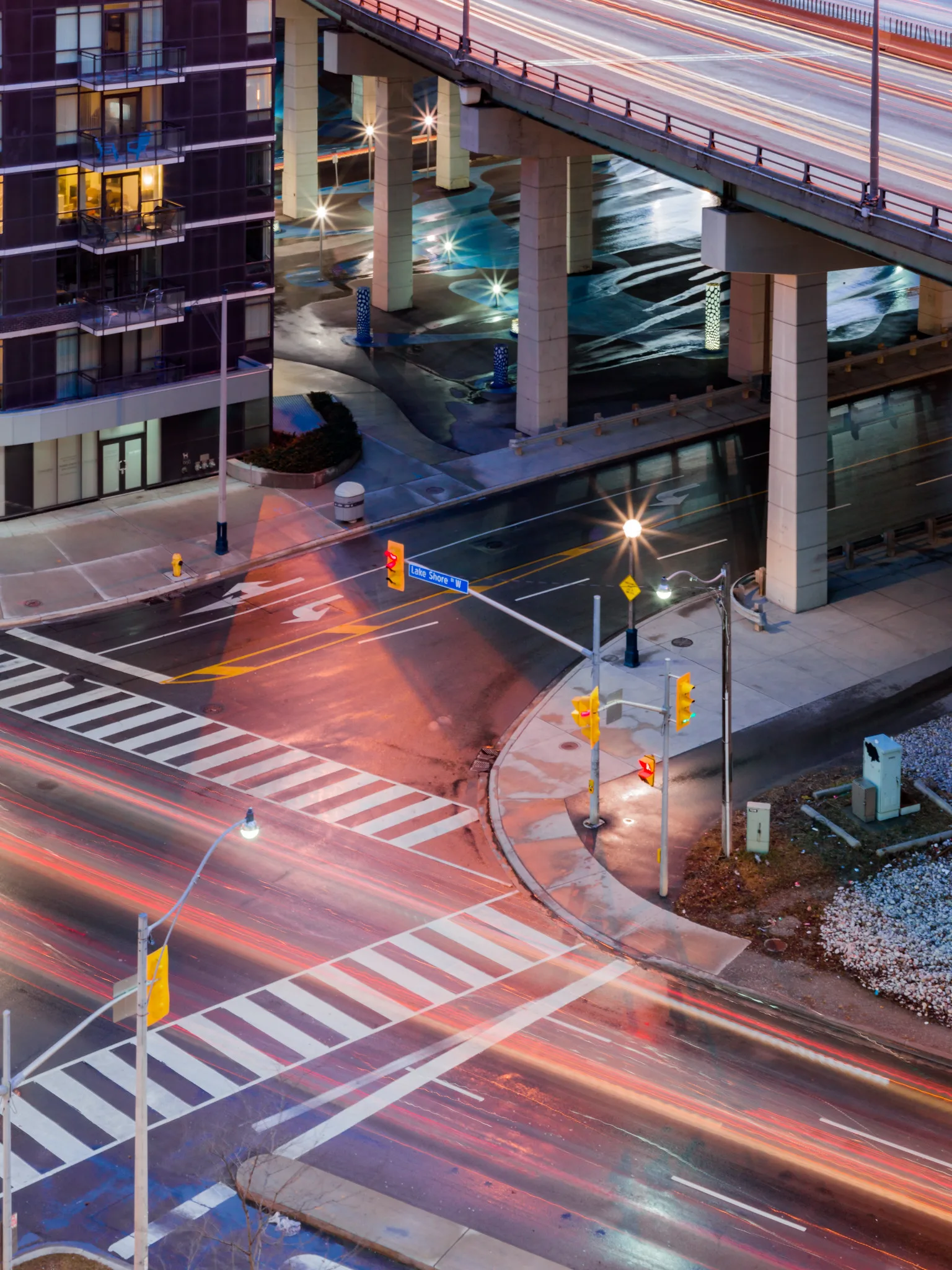 A city intersection at night seen from high above, with red and white streaks of car lights crossing the painted crosswalks and more streaks on an elevated highway behind