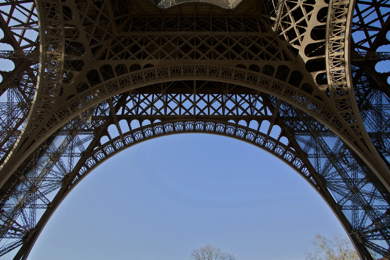 Looking straight up from beneath a huge iron lattice tower, its curved arch and crossed girders framing a clear blue sky