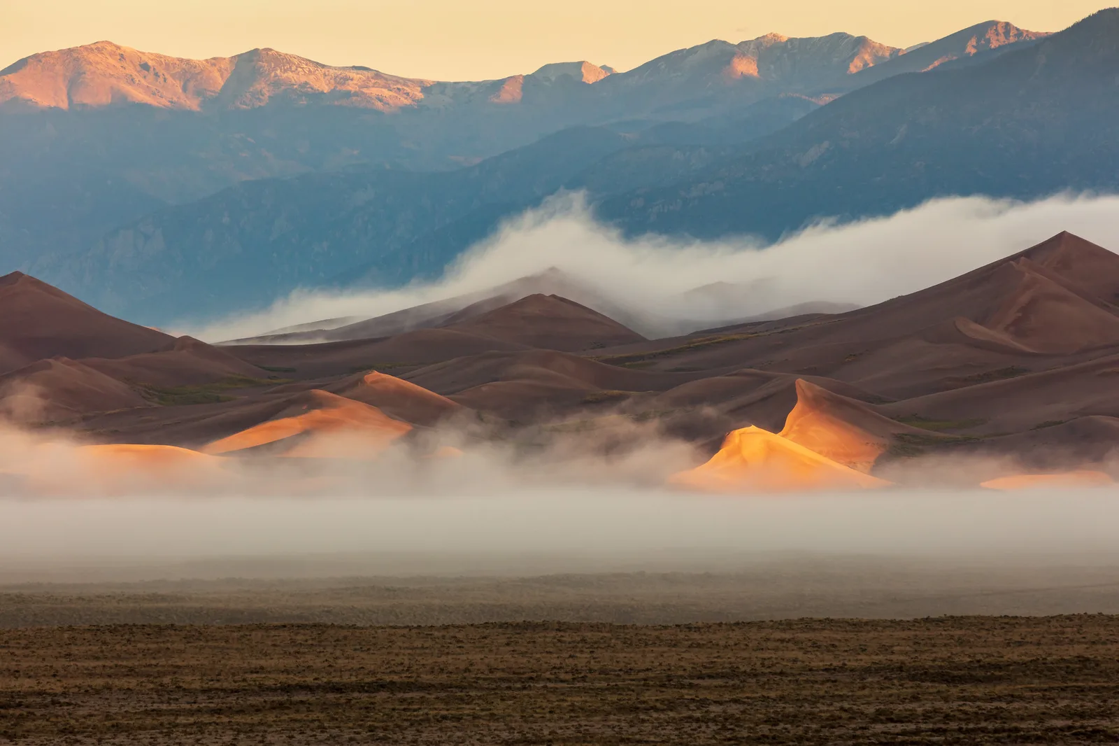 A band of low white cloud drifting across the base of tall sand dunes at sunrise, with blue shadowed mountains behind and a dark plain in front