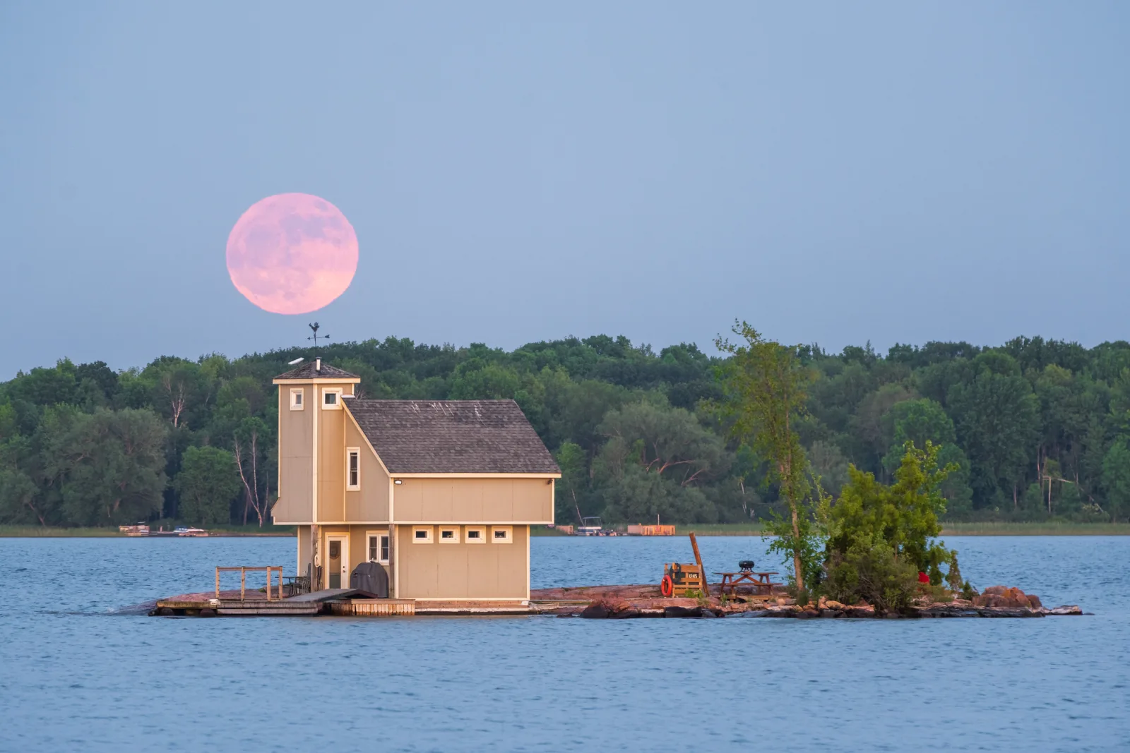 A large pale pink moon low above a line of trees behind a small cottage on a tiny island, with blue water in front and the scene still lit by daylight