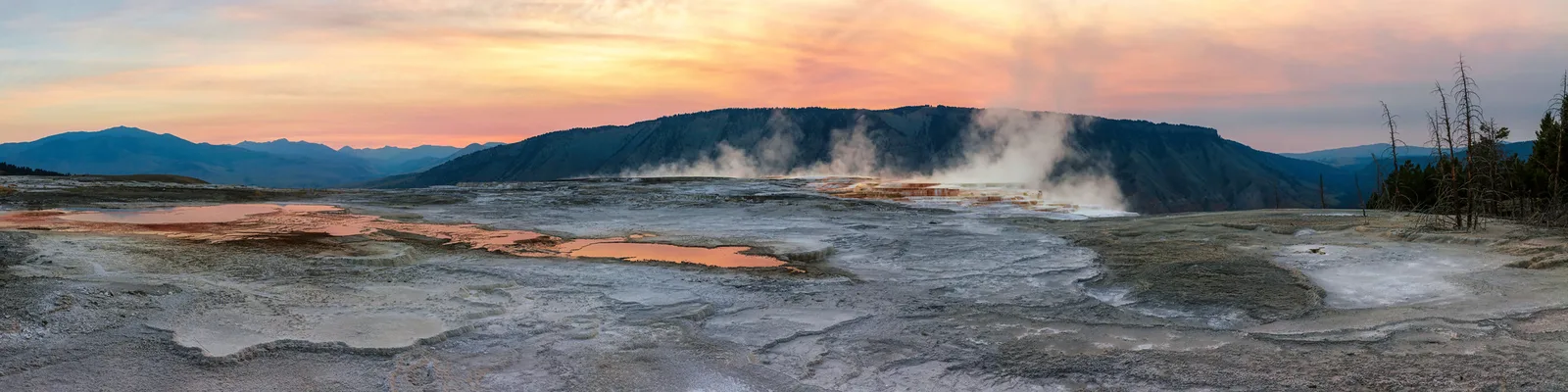 A wide panorama of pale travertine terraces with steam rising from hot springs, dark hills behind and an orange and pink dawn sky
