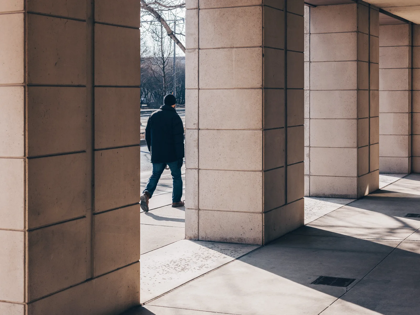 A man in a dark coat walking between tall stone columns in direct sun, with hard-edged black shadows from the columns across the pavement
