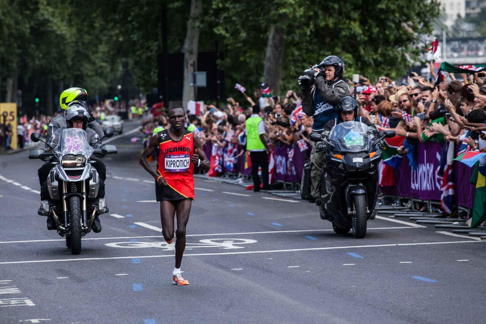A marathon runner in a red vest on a city road between two motorcycle outriders, with a crowd lining the barriers behind