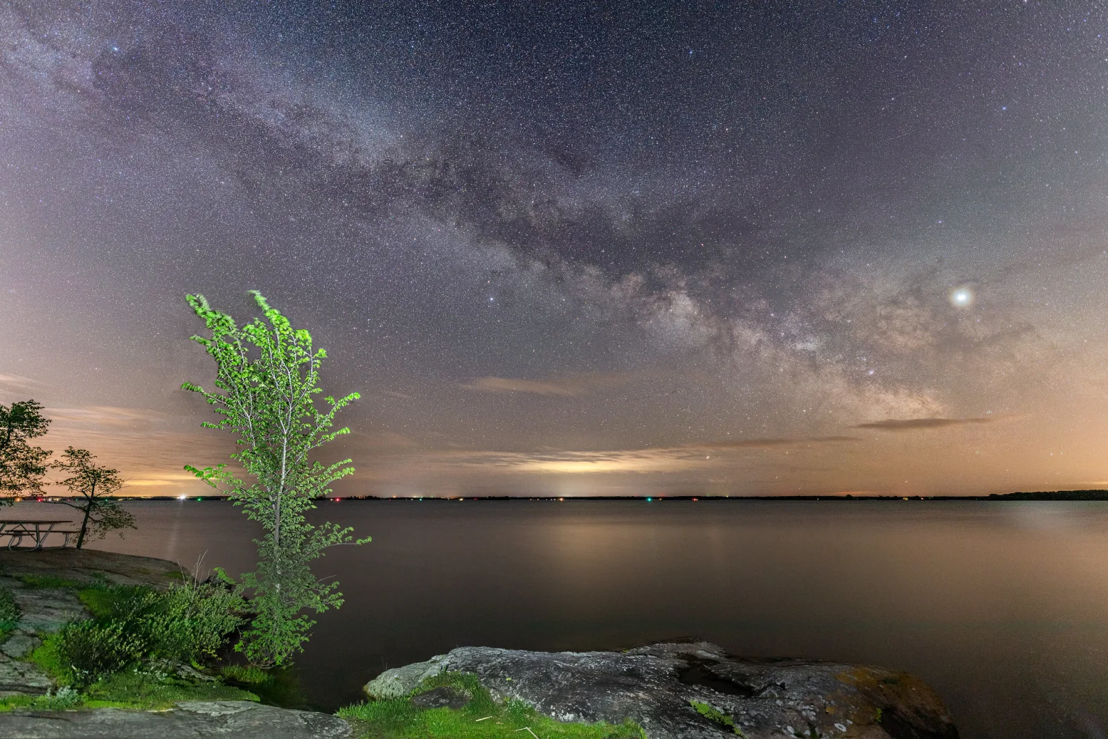 The Milky Way arching over a wide, calm river at night, with a lit green tree and rocks on the shore at left