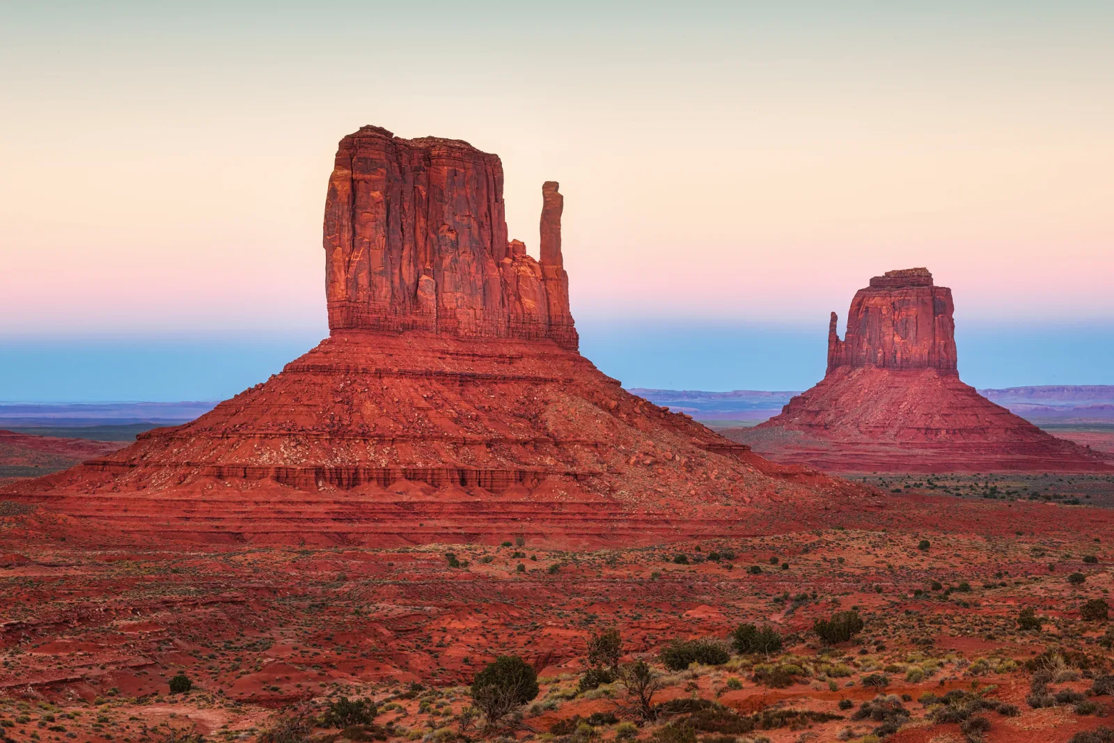Two red sandstone buttes at dusk in a desert valley, glowing deep red under a smooth sky that fades from blue to pale pink