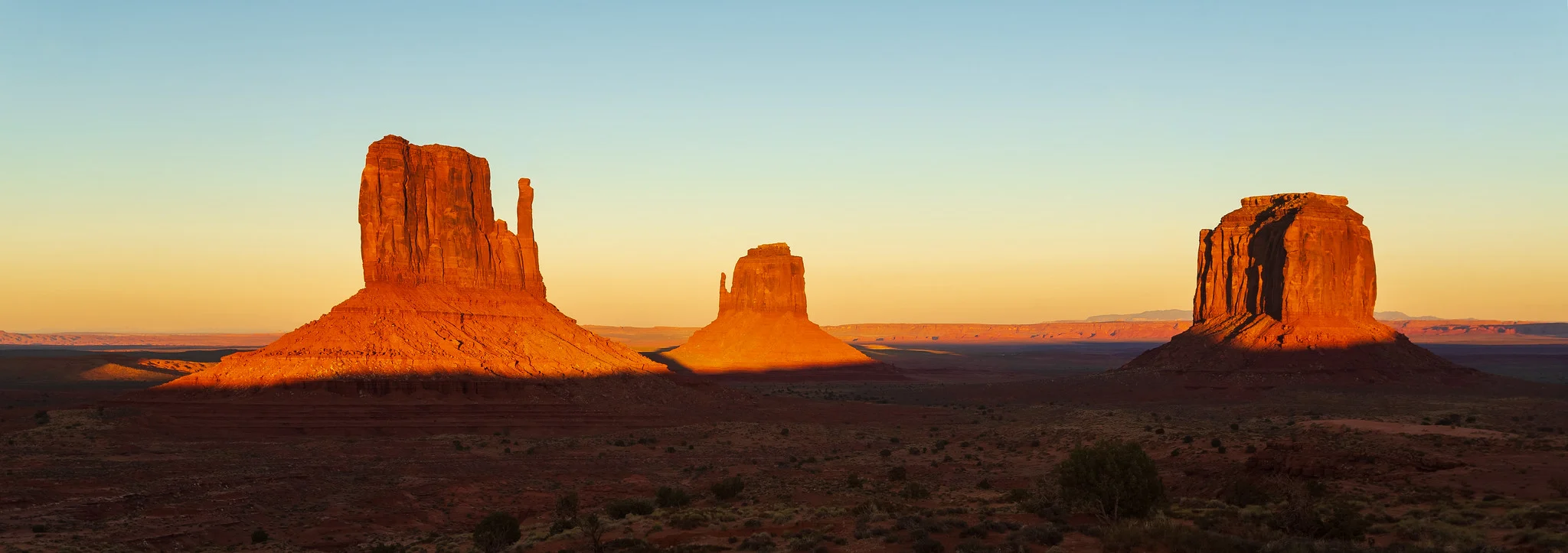 The Mitten buttes of Monument Valley lit orange at sunset against a clear blue sky