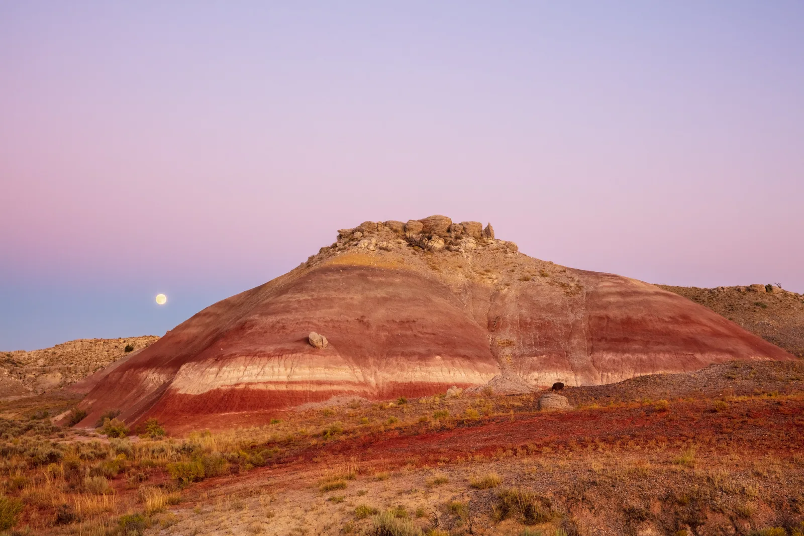 A small moon low in a pink and lavender twilight sky beside a red and cream striped desert hill