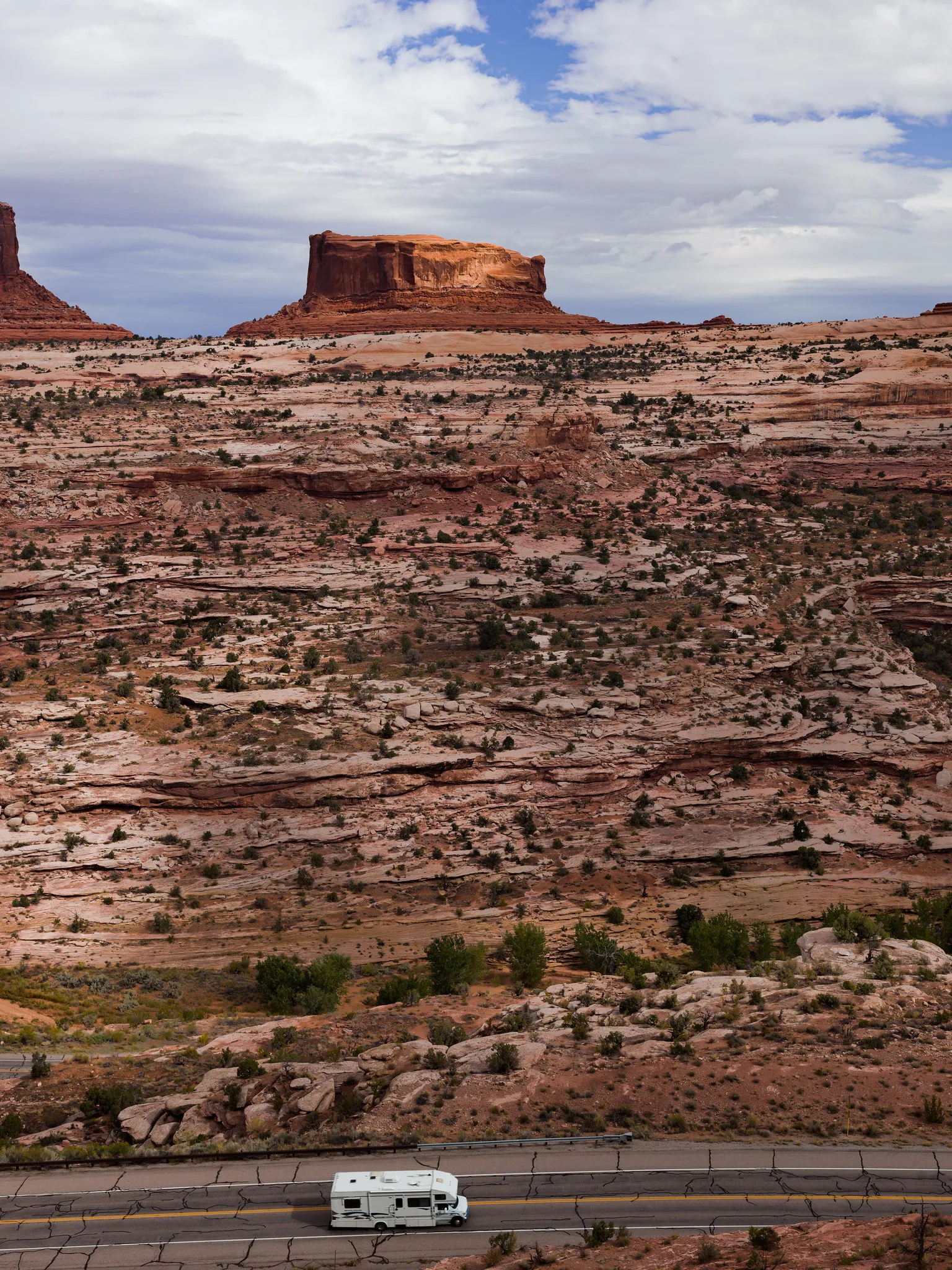 A white motorhome on a road at the bottom of the frame, below a wide expanse of layered red rock ledges and a flat-topped butte under cloud