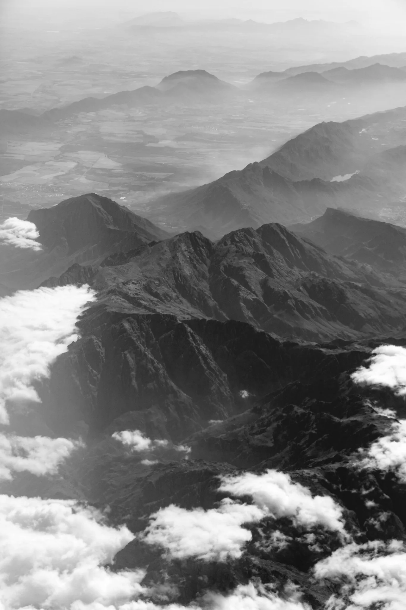 Black and white view from high above of rugged mountain ridges fading into haze, with scattered white clouds below the camera