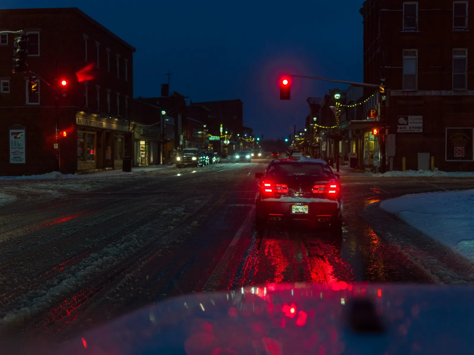A small-town main street at dusk with red traffic lights, a car's glowing tail lights and slushy snow on the road