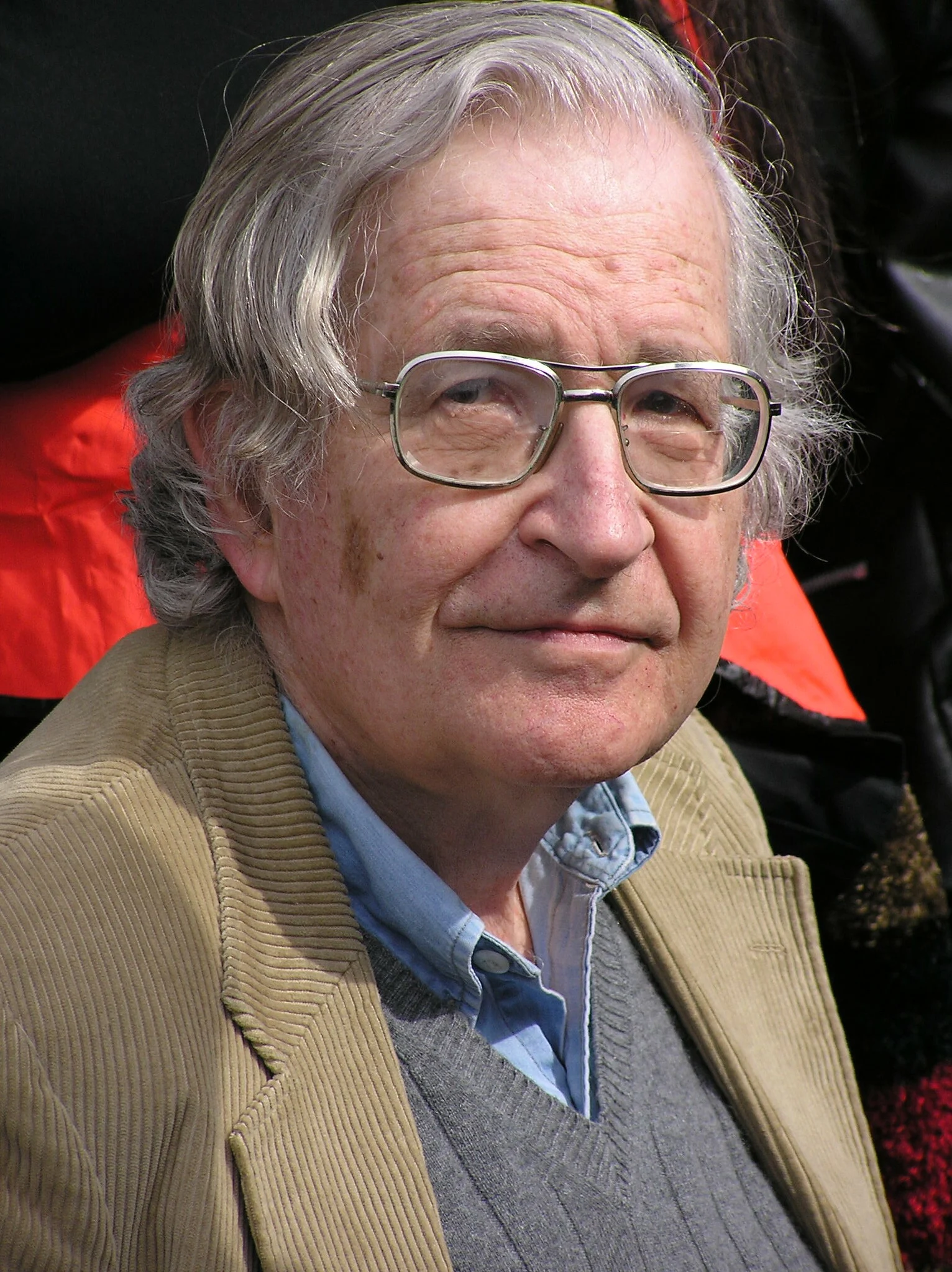 A documentary-style outdoor portrait of an older man with gray hair and glasses, wearing a corduroy jacket
