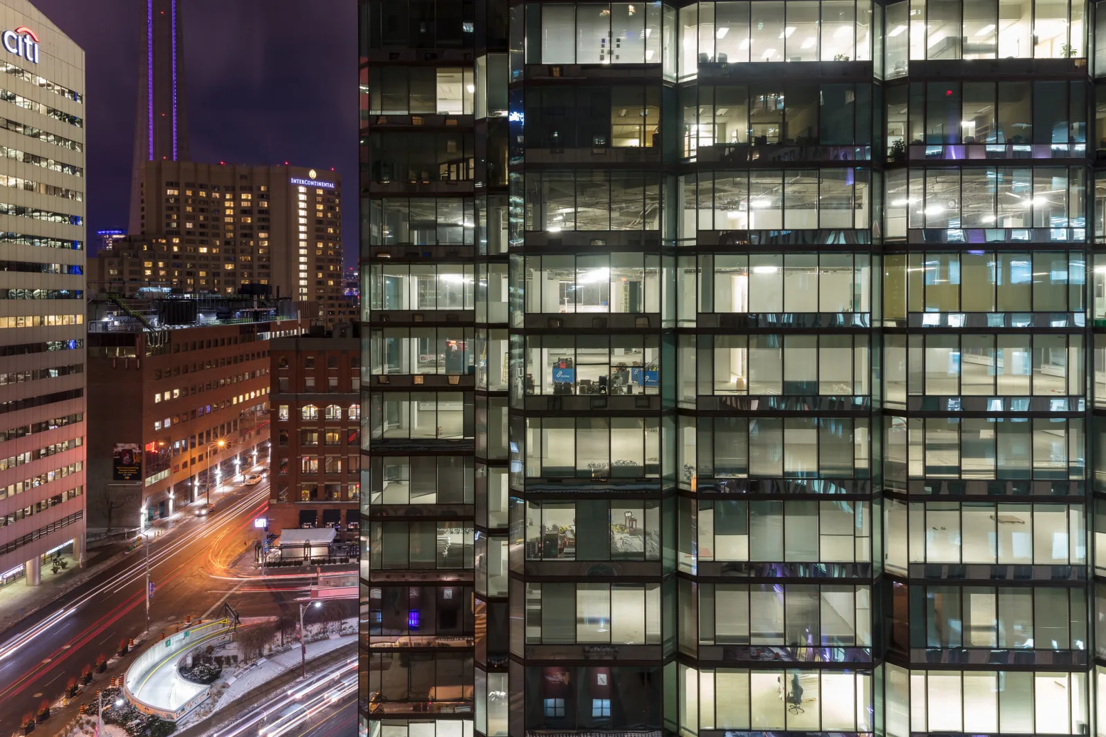 A glass office tower at night with dozens of lit windows beside a street with light trails and other towers
