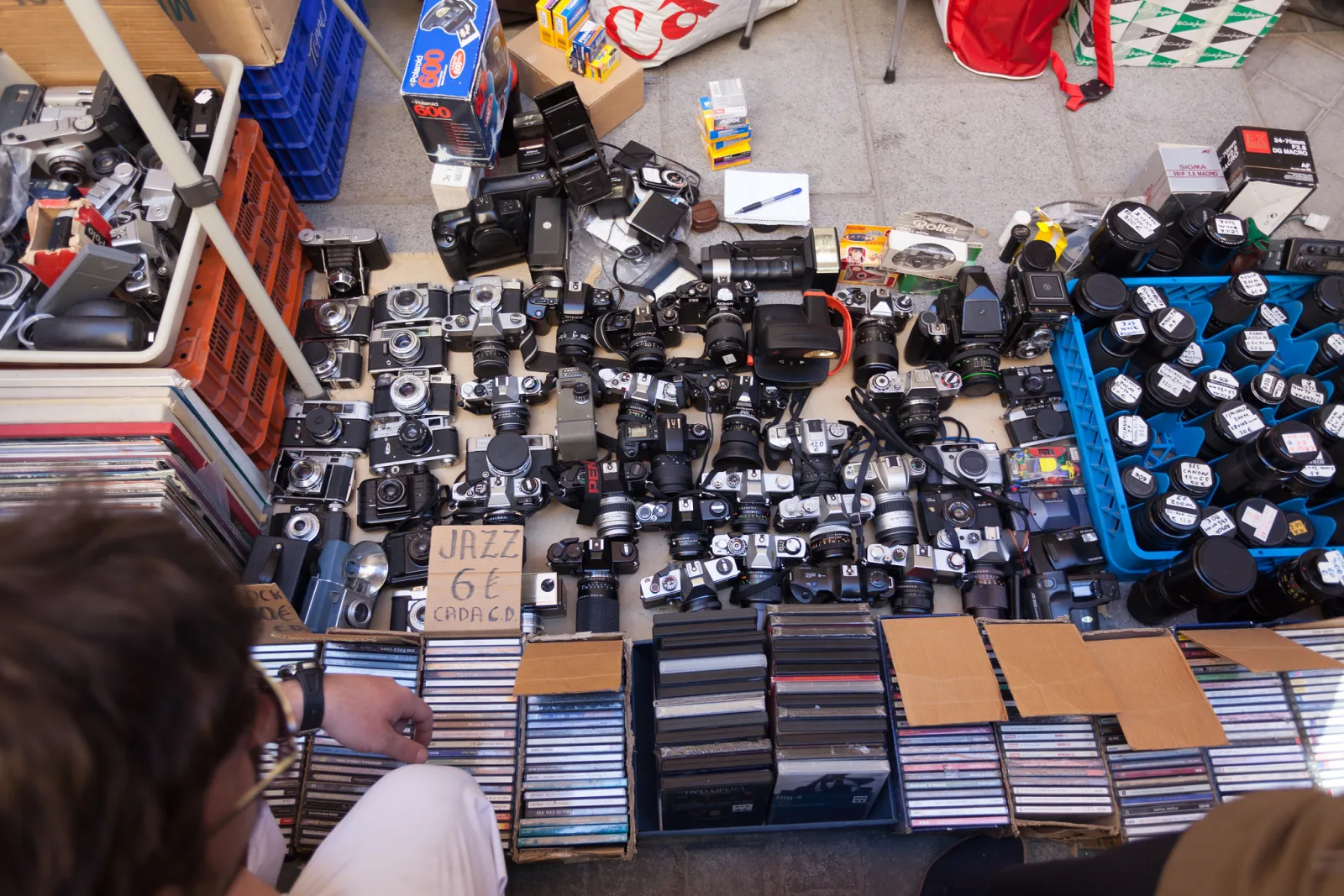 A flea market table covered with dozens of old film cameras, lenses and boxes, with shoppers' hands at the edge of the frame