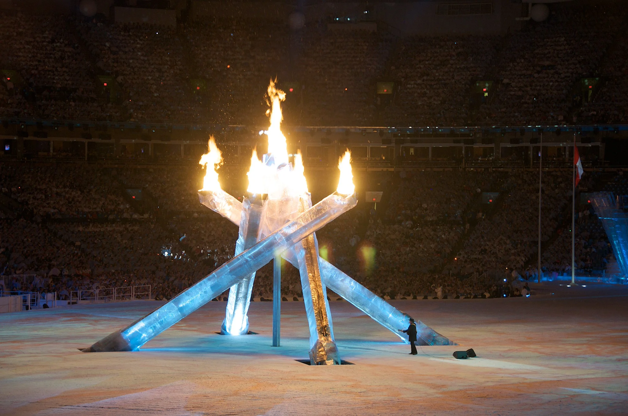The Olympic cauldron burning brightly at the center of a dark stadium, light falling off rapidly toward the seating
