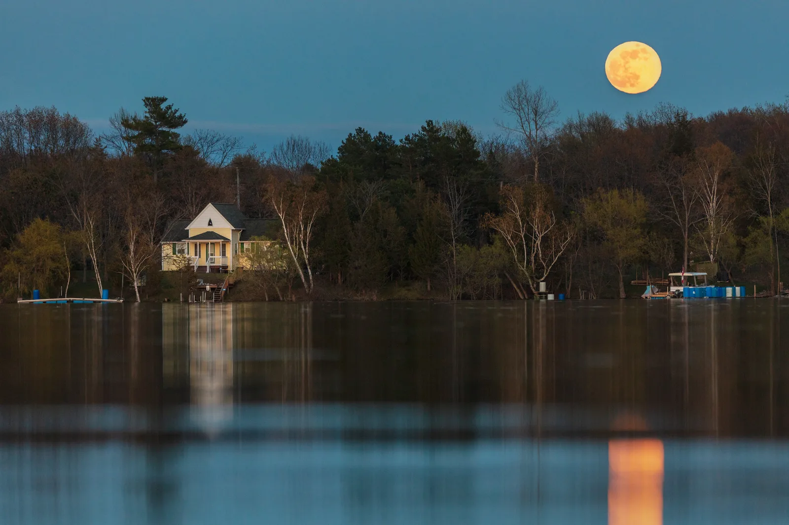 An orange full moon low in a blue dusk sky above bare trees and a pale yellow cottage on a lake shore, with the moon reflected in the dark water