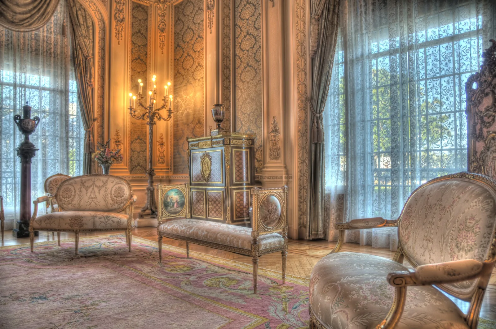 An ornate historic room with gilded walls, patterned rug, upholstered chairs and candelabras, lit by tall bright windows with lace curtains