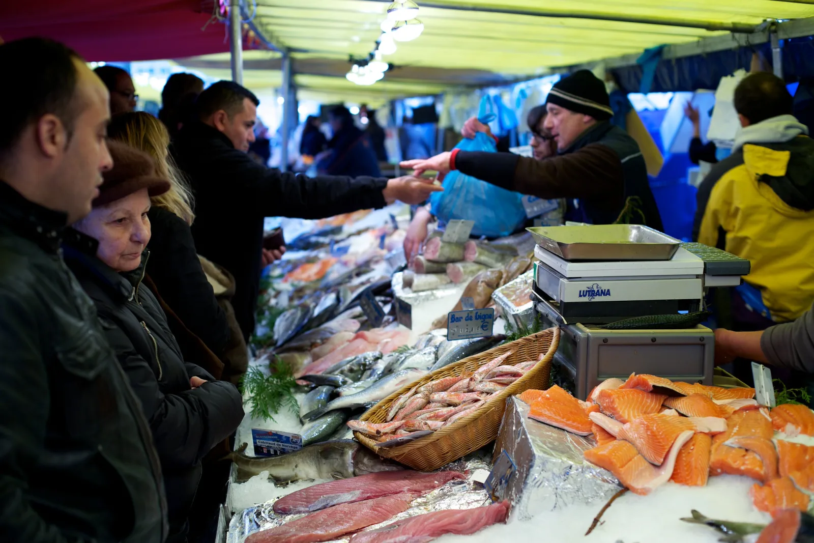 Customers leaning over a fish stall under a green awning while a fishmonger passes a bag across the counter, with salmon steaks and whole fish on ice in front