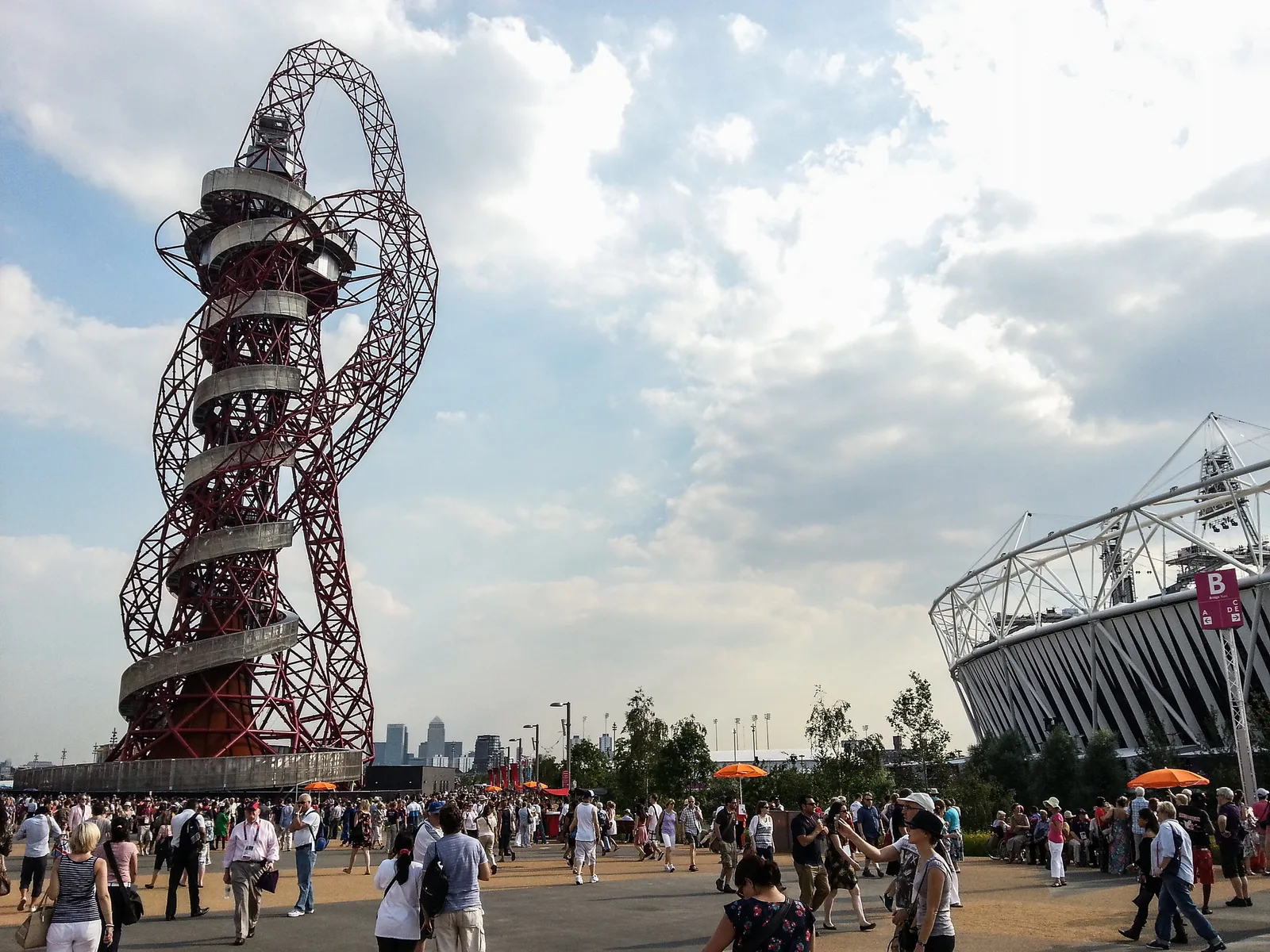 A red looping steel observation tower beside a stadium under a bright cloudy sky, with a crowd of people walking across the plaza in front