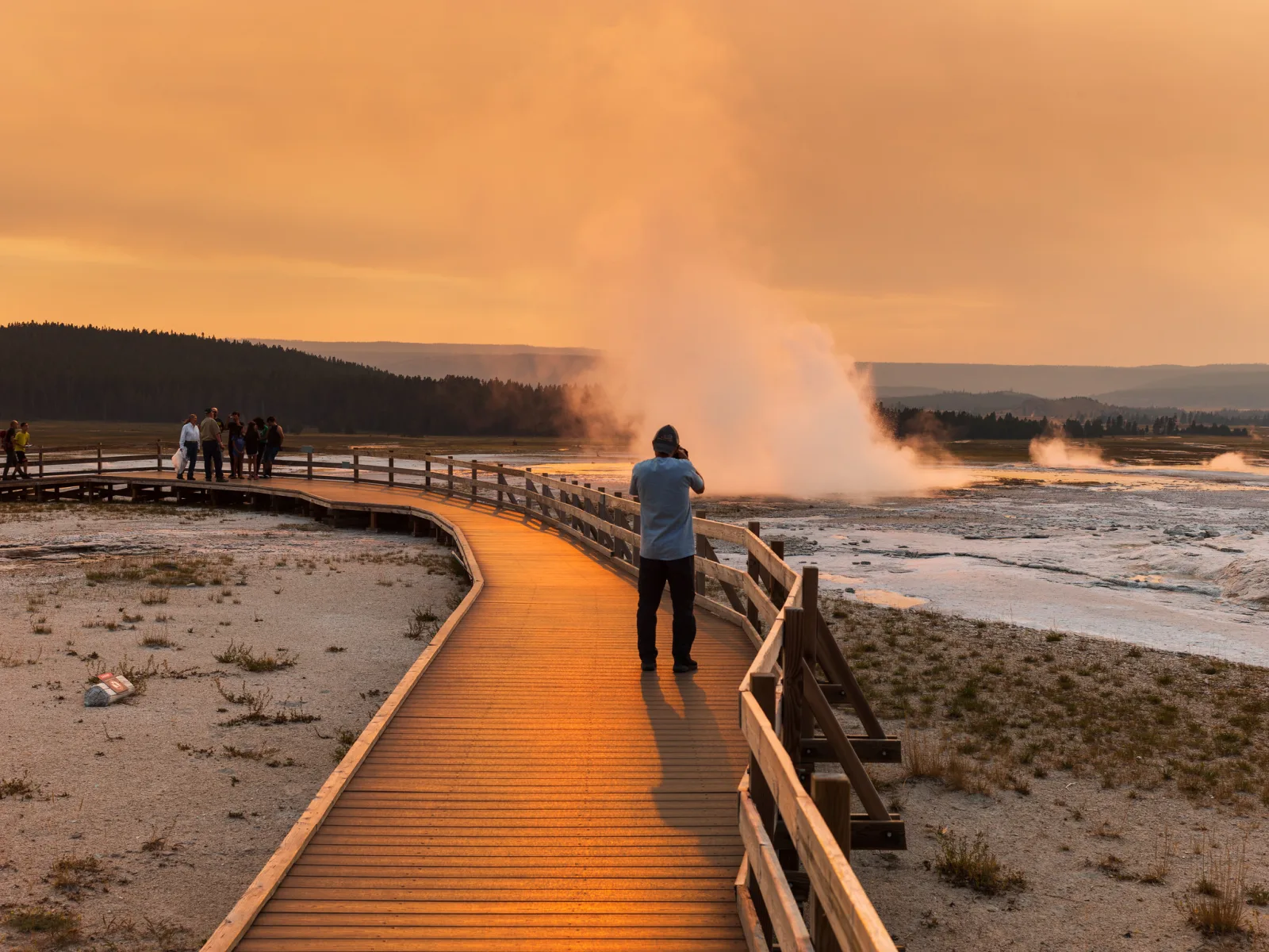 A photographer standing on a wooden boardwalk shooting a steaming geyser in orange sunrise light, with other visitors further along the railing