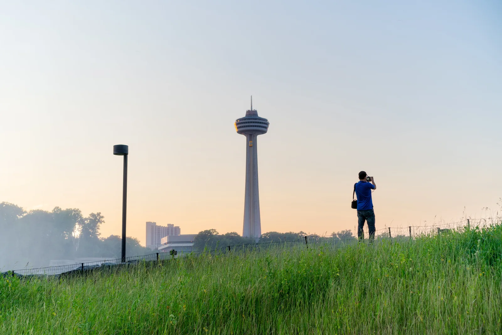 A lone person standing on a grassy rise at dusk holding a camera up to their eye, with an observation tower and a lamp post against a pale sky