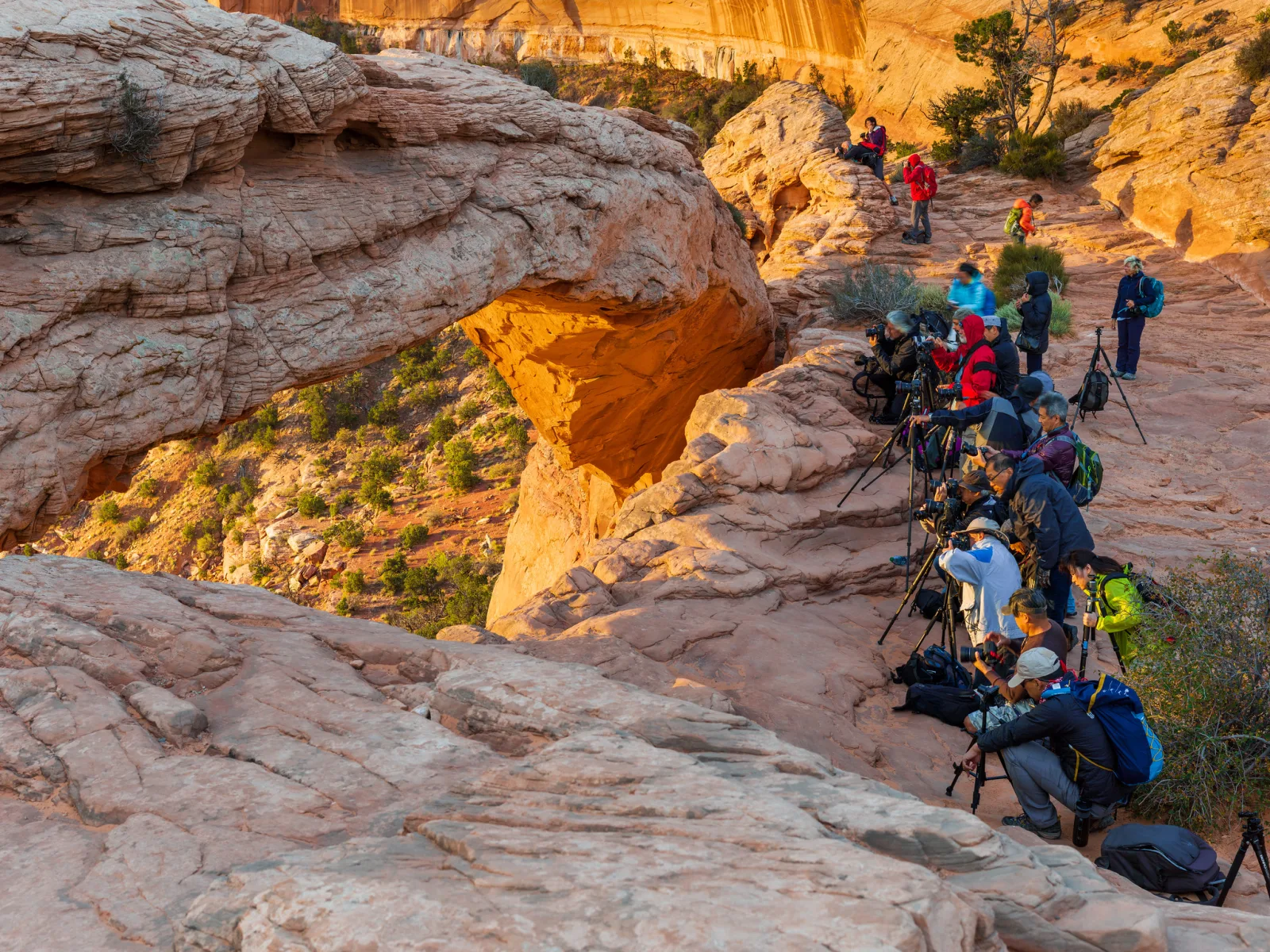 A line of photographers with cameras on tripods crowded along a sandstone ledge beside a rock arch