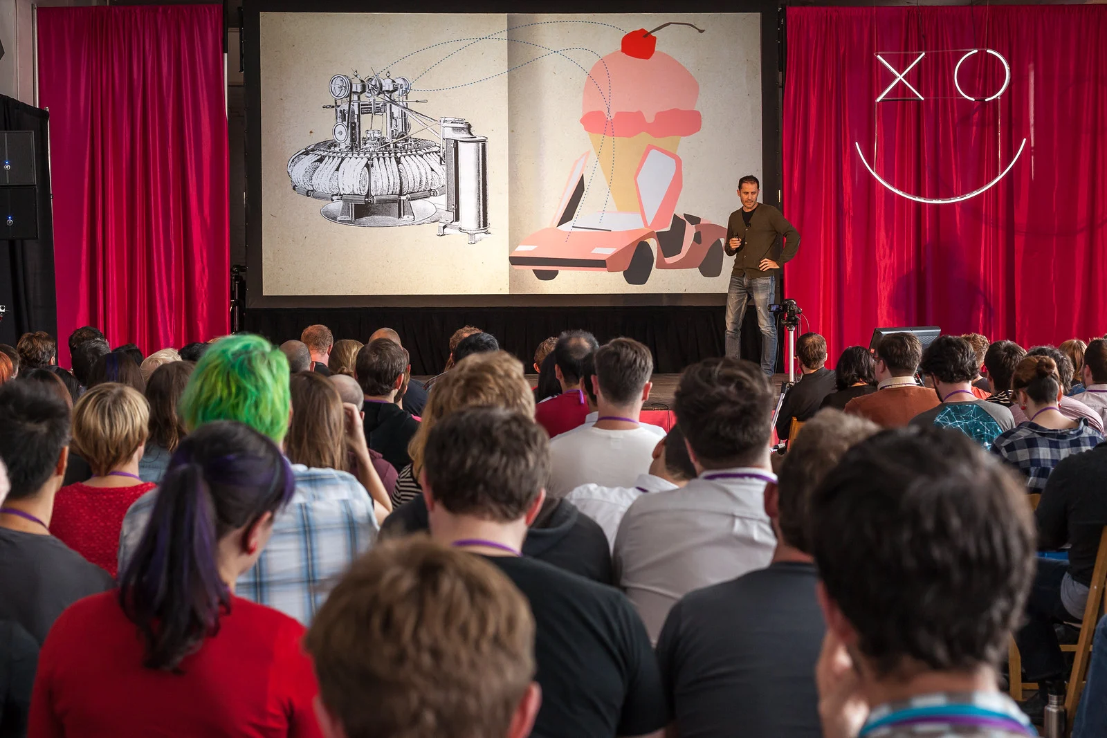 A speaker presenting on stage to a full audience at a conference, photographed from the crowd