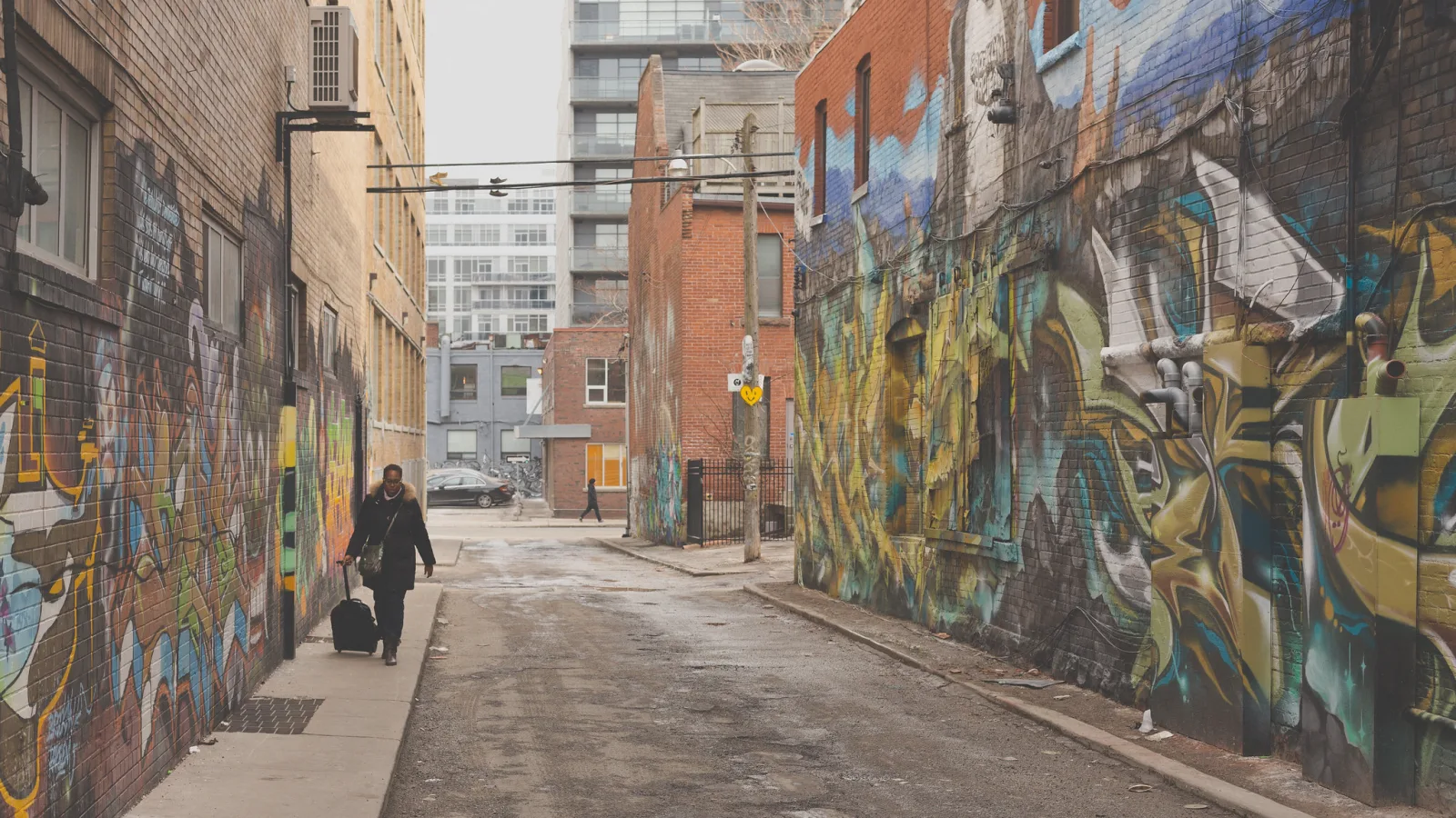 A person pulling a wheeled suitcase down a narrow alley whose brick walls are covered in large colorful painted murals, with apartment buildings beyond