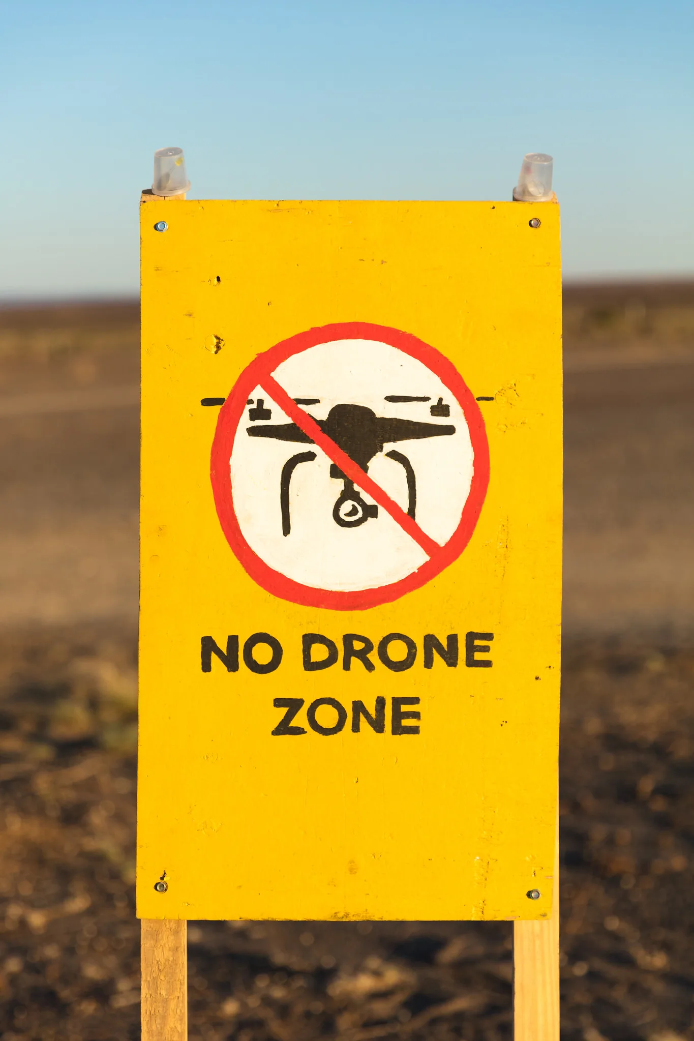 A yellow sign on wooden posts reading No Drone Zone, with a drone symbol crossed out in a red circle, standing in a dry field