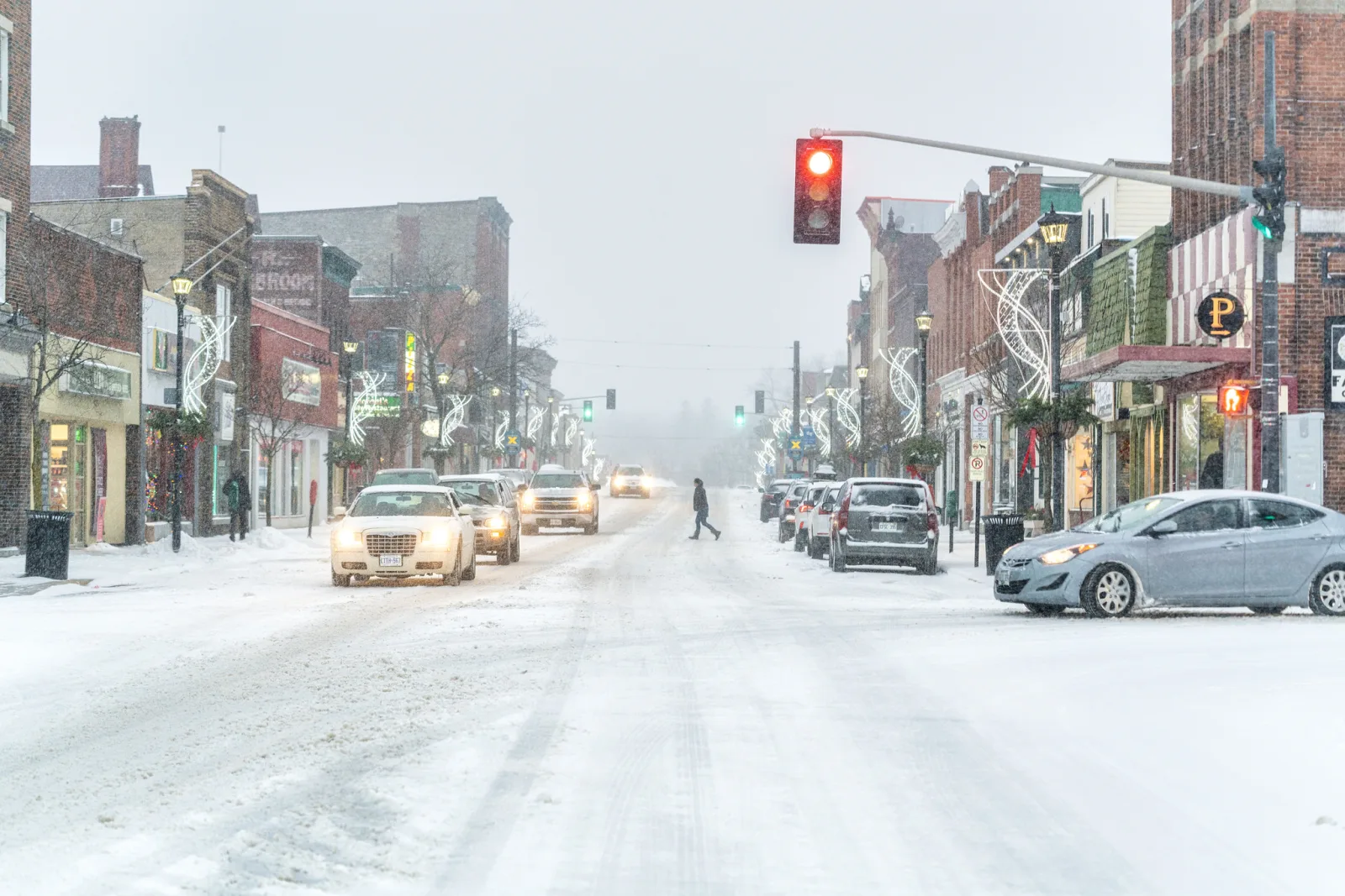 A snowy main street in a small town with shopfronts on both sides, cars driving and parked, a red traffic light and a person crossing the road in the distance
