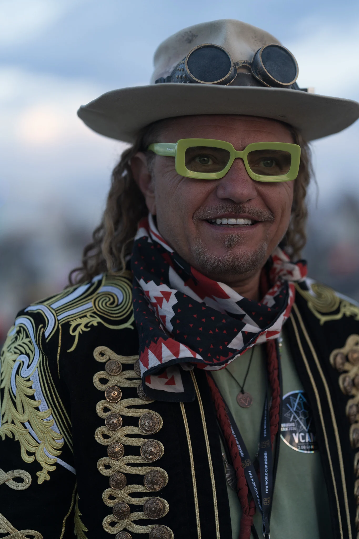 A close portrait of a man in a wide-brim hat, green sunglasses and an embroidered gold jacket, with the background thrown out of focus