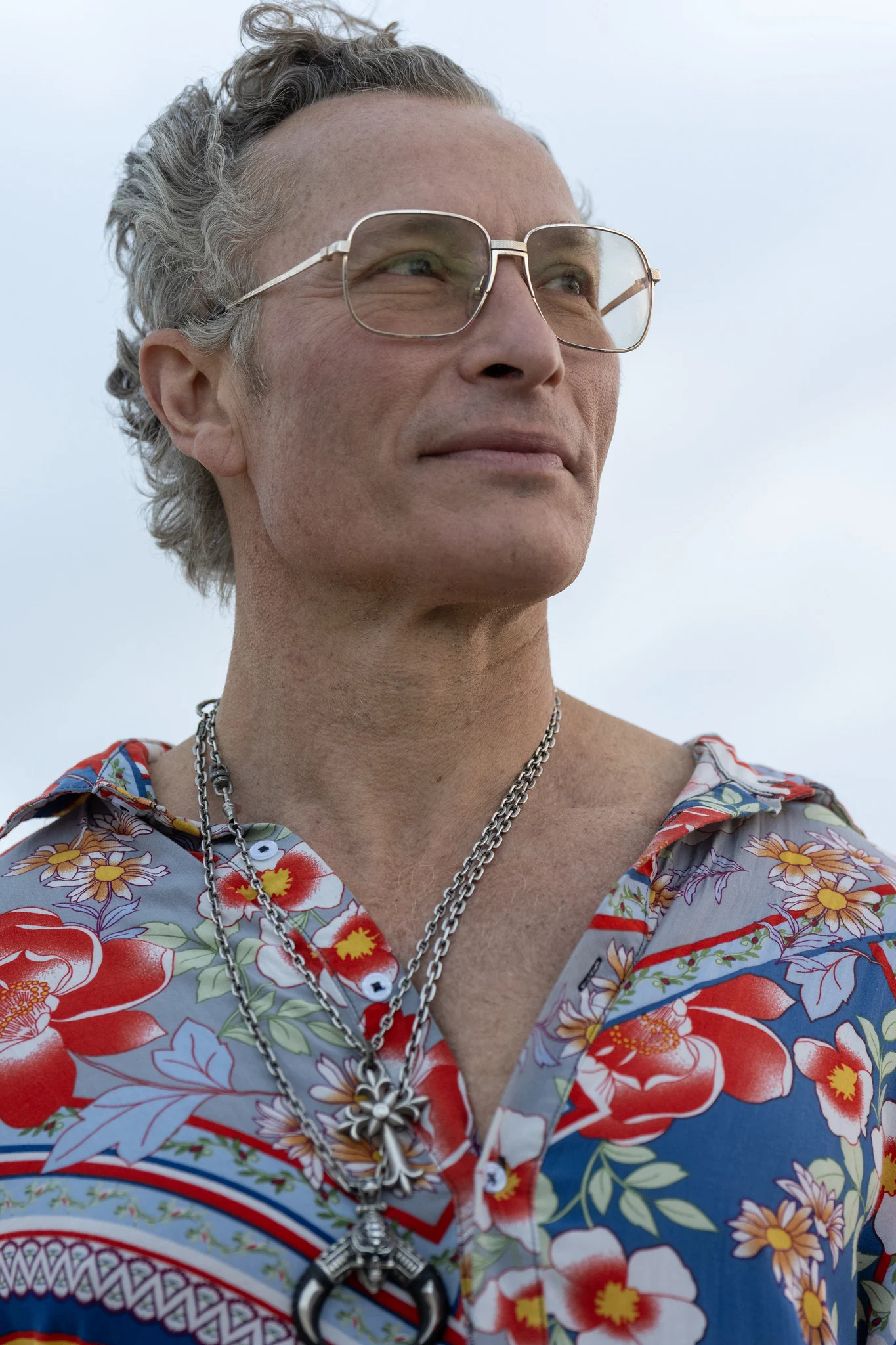 A close portrait of a man in wire glasses and a floral shirt looking off camera in soft, overcast morning light