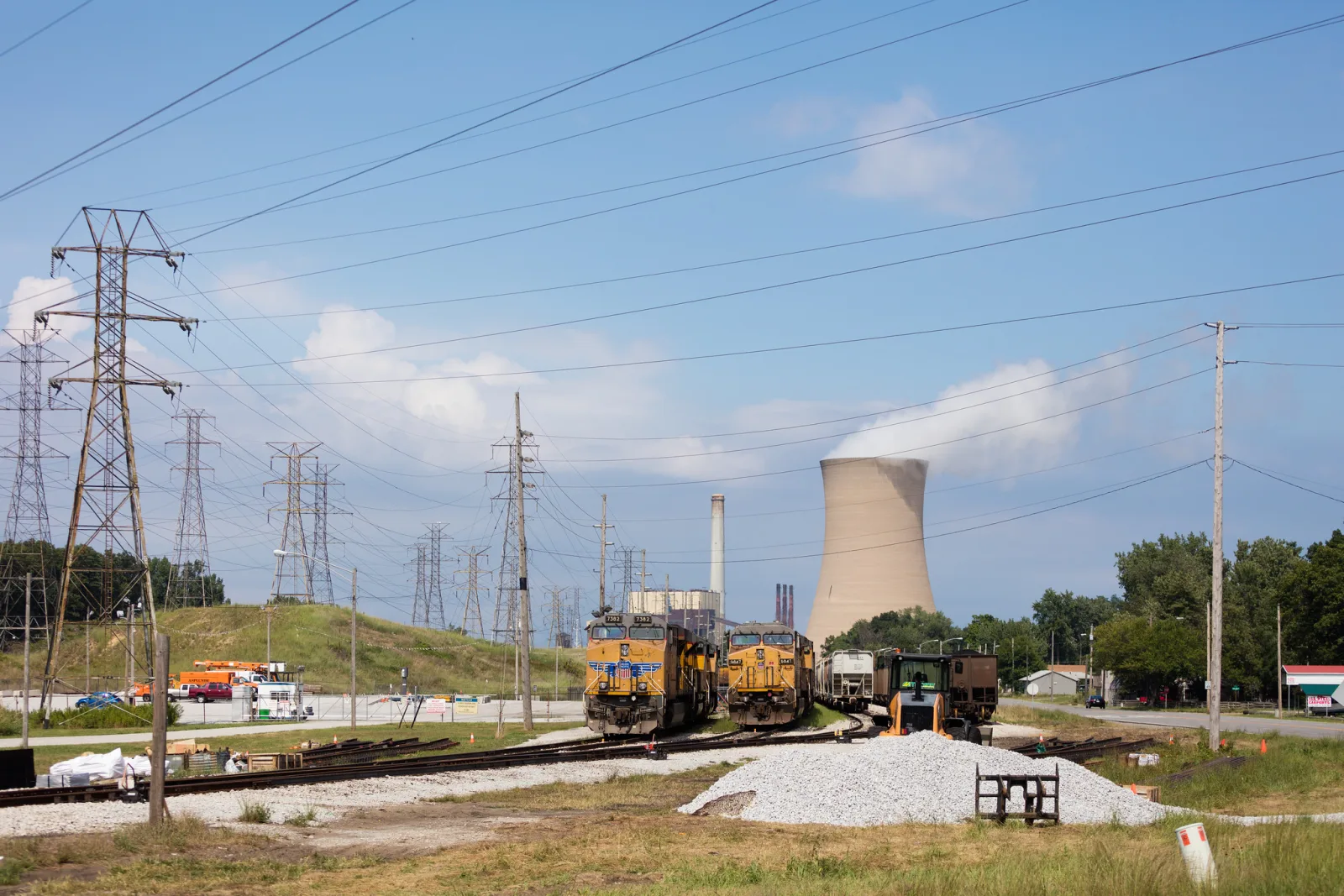 A rail yard with yellow locomotives, gravel piles and transmission towers, with a steaming cooling tower behind under a blue sky