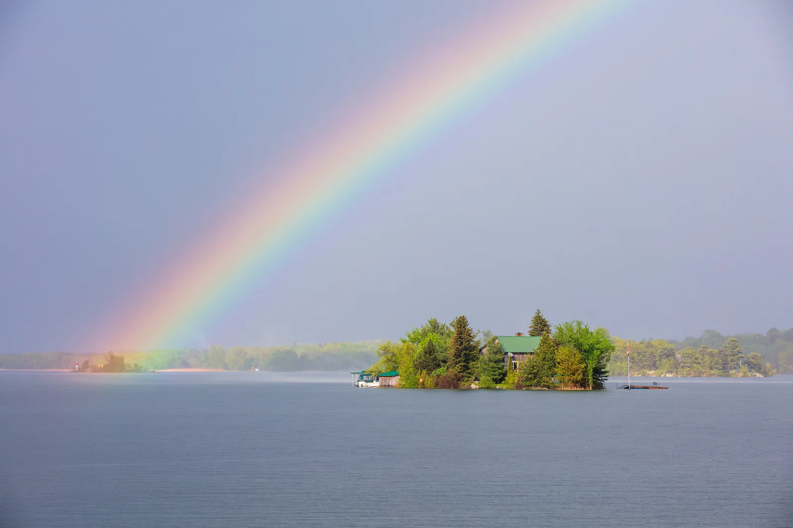 A bright rainbow arcing down to a small wooded island on a gray lake under a stormy sky
