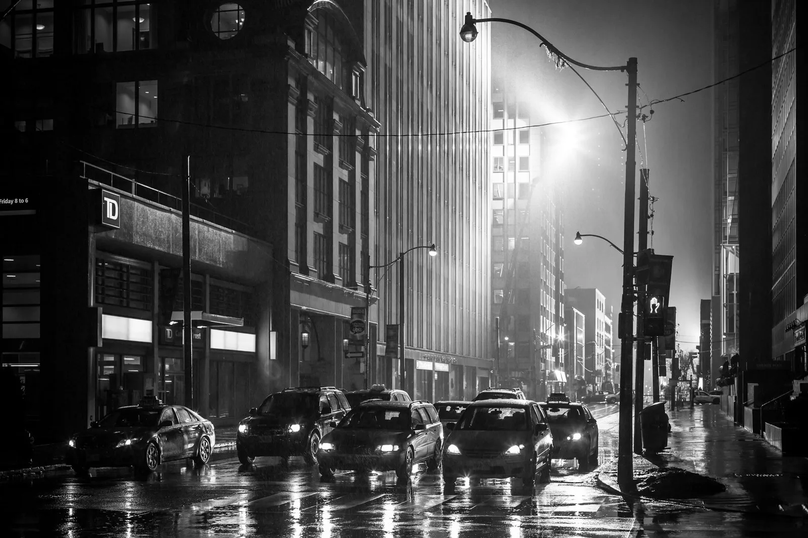 Black and white night street between tall buildings, with wet pavement reflecting car headlights and streetlights glowing in the rain