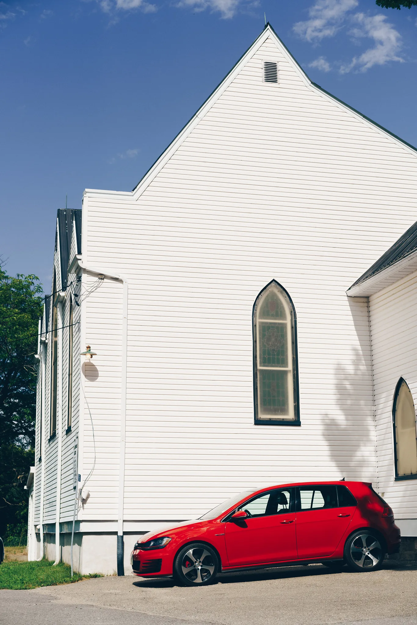A white wooden church with a pointed gable and arched windows against blue sky, a red hatchback parked beside it