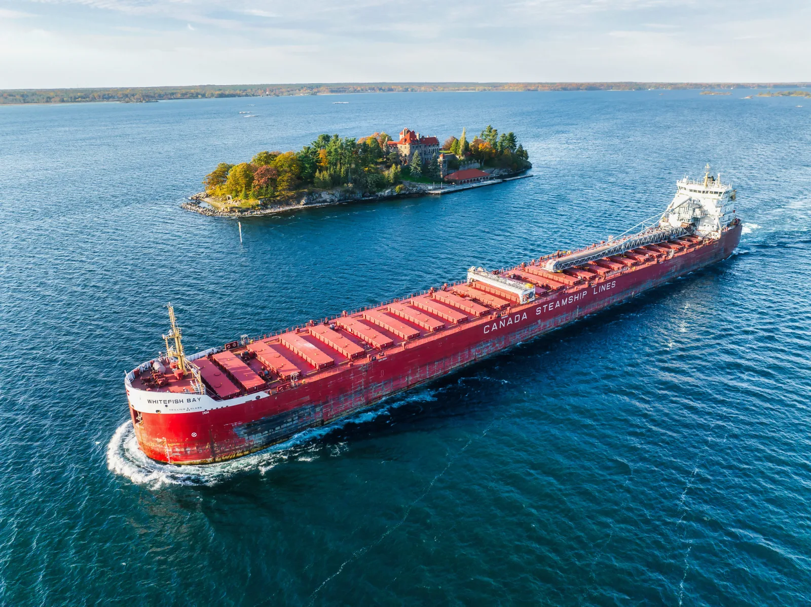 Aerial view of a long red cargo ship moving through deep blue water past a small wooded island
