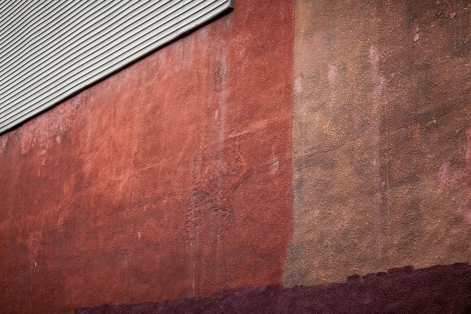 A rough red and rust coloured painted wall filling the frame, with a strip of grey corrugated metal in the top left corner