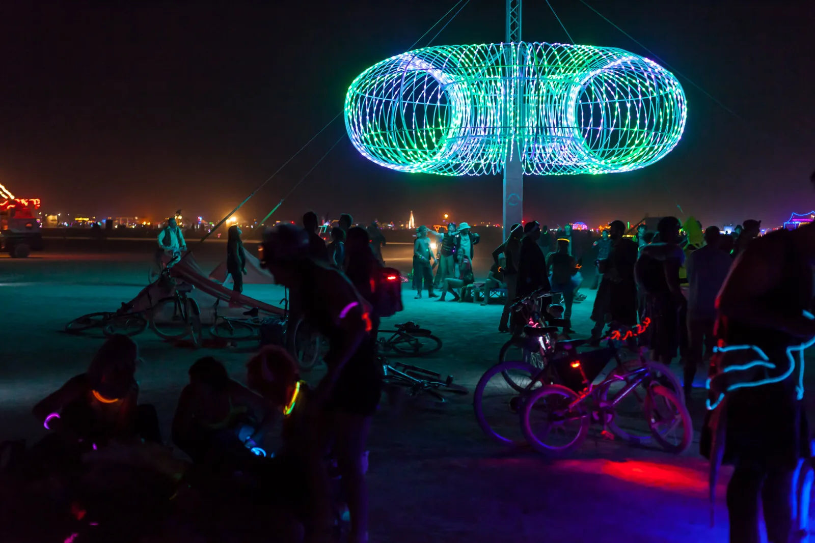 A large ring-shaped sculpture made of glowing green and blue LED lines at night, with people, bicycles and small coloured lights in the dark foreground