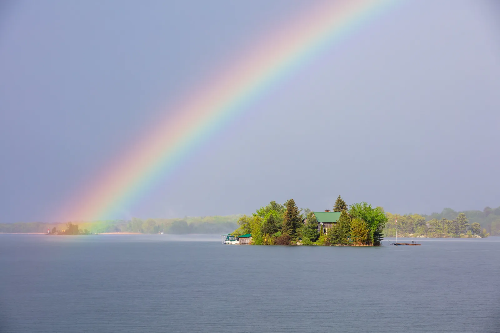 A bright rainbow arcing down to a small wooded island in a calm grey lake under an overcast sky