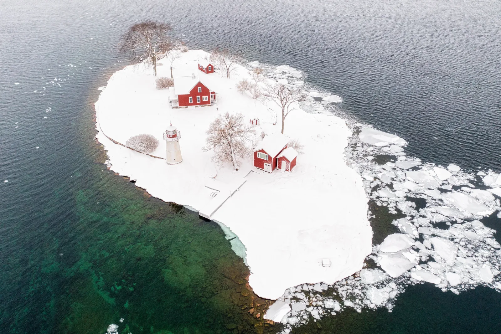Aerial view of a small snow-covered island with a white lighthouse and red buildings, surrounded by dark water and floating ice